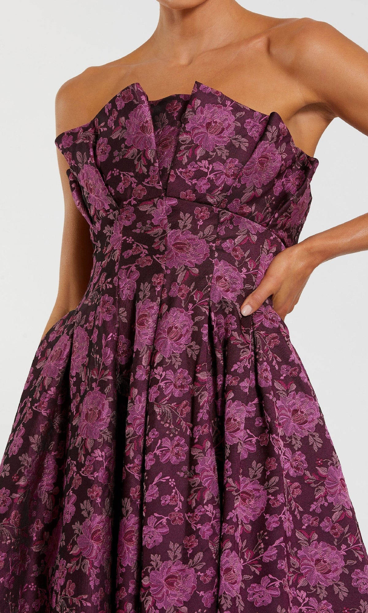 A person poses with one hand on their hip in the Mac Duggal 56159 Strapless Wedding Guest Dress, featuring a dark purple hue, pink floral pattern, and pleated bodice detailing.